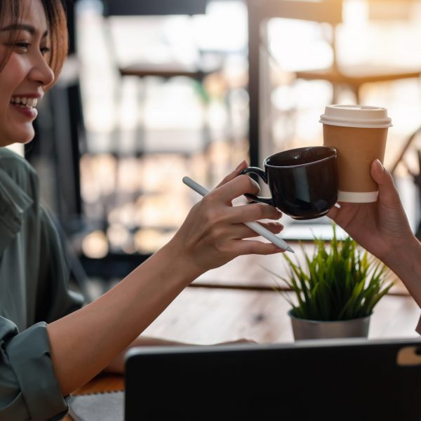 Happy working Asian woman hold coffee cups and cheers at café.